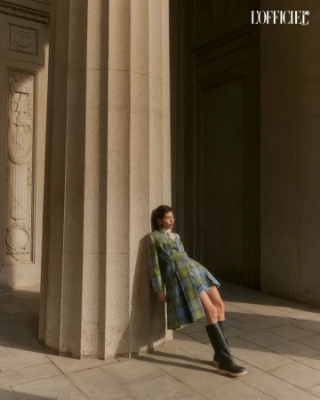 A woman poses in a plaid dress and black boots in front of a column, bathed in sunlight, with the L'Officiel logo visible.