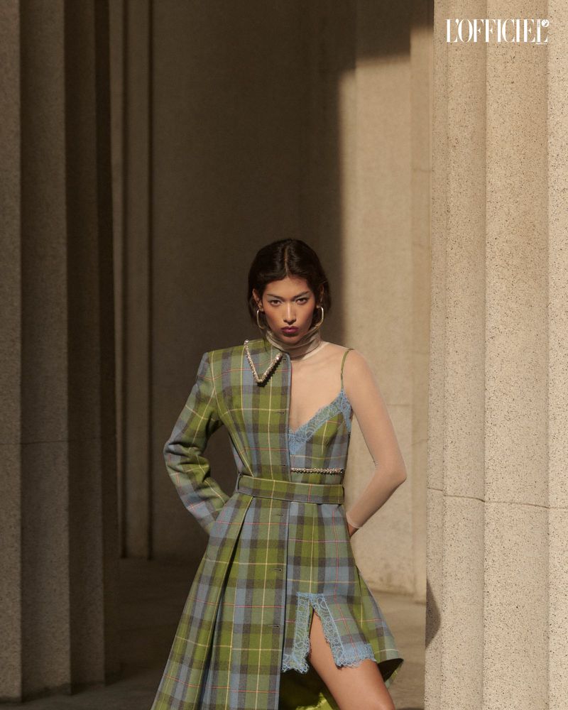 A stylish woman in a plaid dress stands confidently in front of stone columns, accessorized with hoop earrings and neutral makeup.