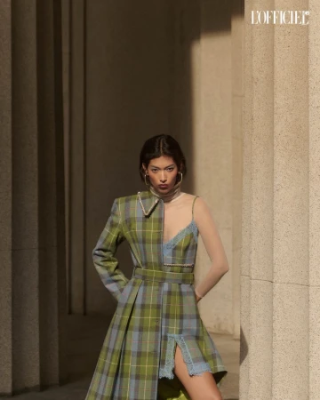 A stylish woman in a plaid dress stands confidently in front of stone columns, accessorized with hoop earrings and neutral makeup.