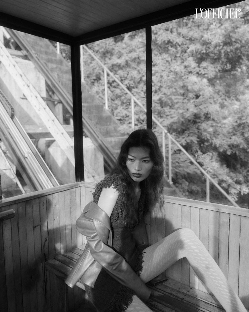 Black and white fashion shot of an Asian woman in a wooden structure, wearing a dress, jacket, and patterned leggings with trees visible.