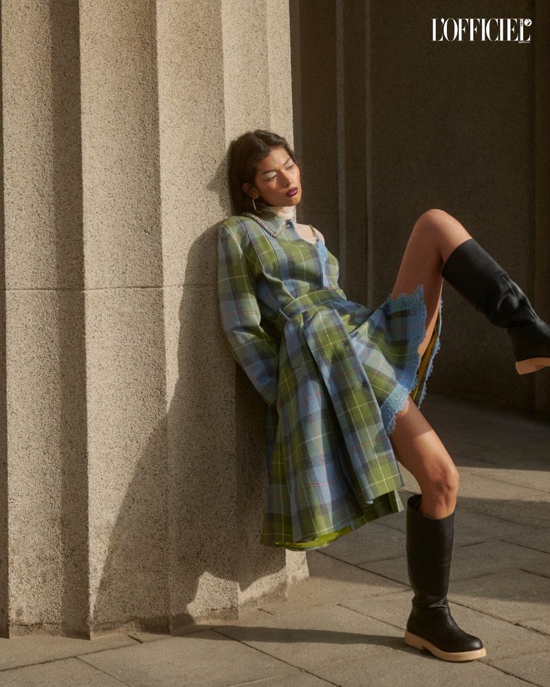 A woman leans against a stone column wearing a plaid dress and black boots, bathed in soft sunlight. The style is editorial fashion.