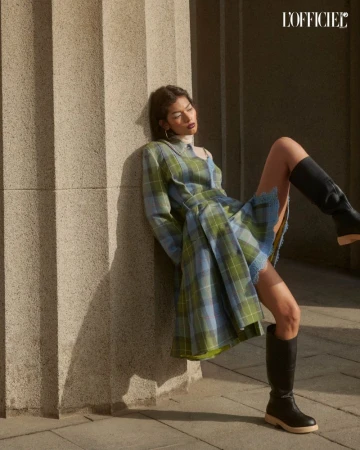 A woman leans against a stone column wearing a plaid dress and black boots, bathed in soft sunlight. The style is editorial fashion.