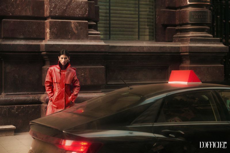 Brunette woman in a red trench coat stands next to a black taxi cab with a red light in front of a stone building.