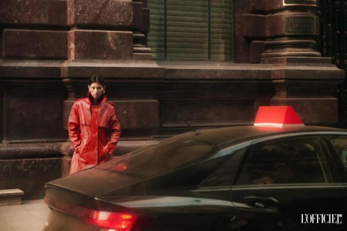 Brunette woman in a red trench coat stands next to a black taxi cab with a red light in front of a stone building.