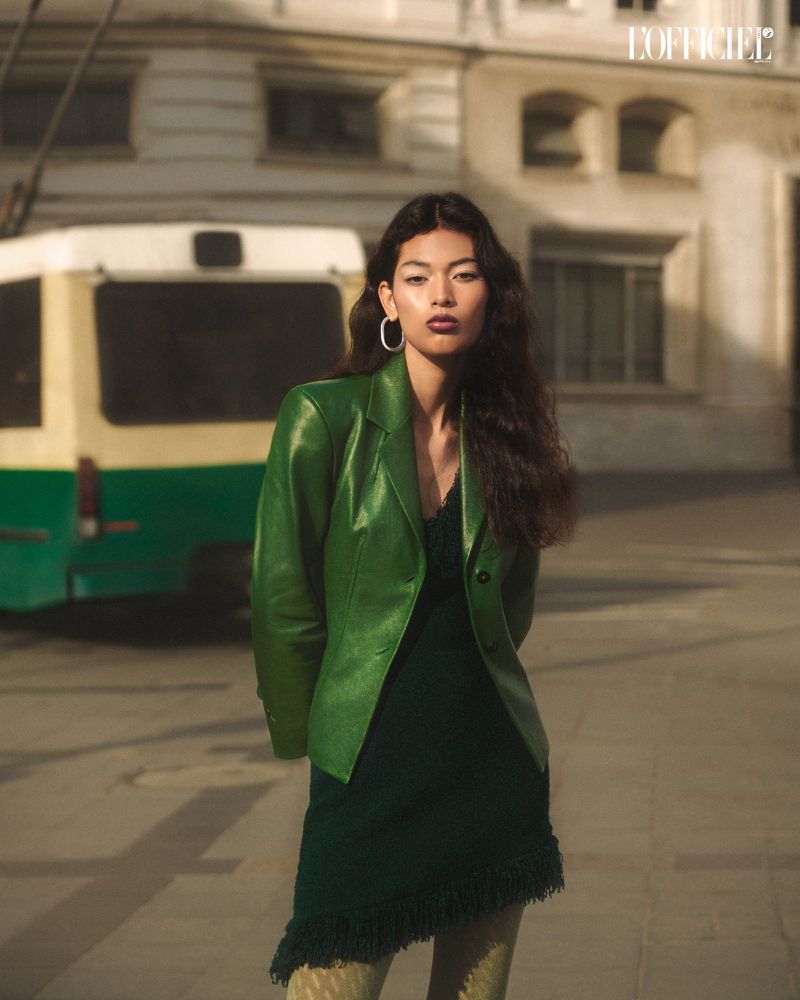 A confident woman in a green jacket and dress stands in an urban setting with a bus and buildings in the background, fashionable.