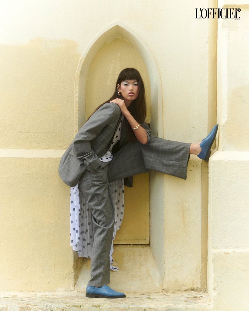 Fashion editorial shot of an Asian woman in a grey suit and blue shoes posing in an arched doorway with the L'Officiel magazine logo.