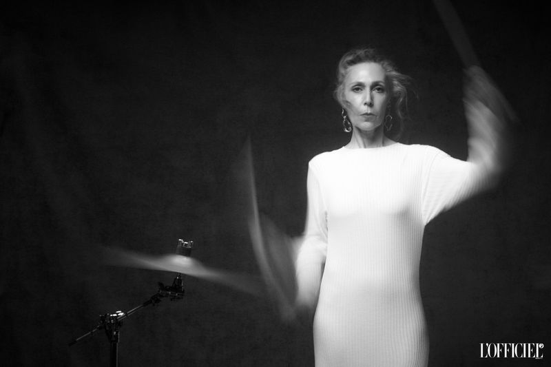 Striking black-and-white portrait of a woman drumming with a cymbal, wearing a white dress and dangling earrings, set against a dark backdrop.
