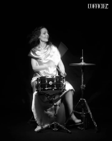 Adult woman playing a drum set in a photo studio against a black background, wearing a draped textile and illuminated with studio lighting.