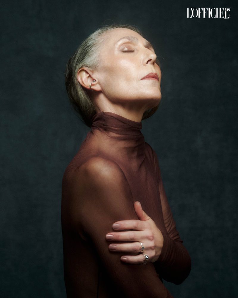 Portrait of a mature adult woman with gray hair wearing a brown turtleneck with arms crossed in front of a dark gray background.
