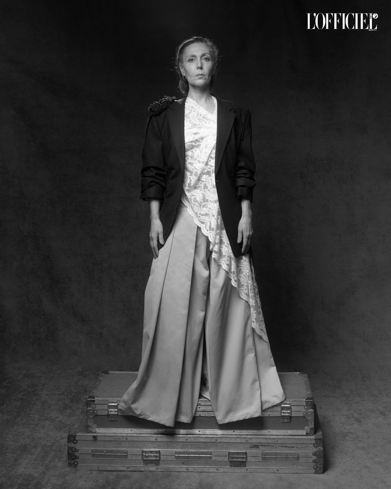 Black and white portrait of Juanita Parra standing on stacked vintage trunks in an oversized blazer and pleated wide-leg trousers.