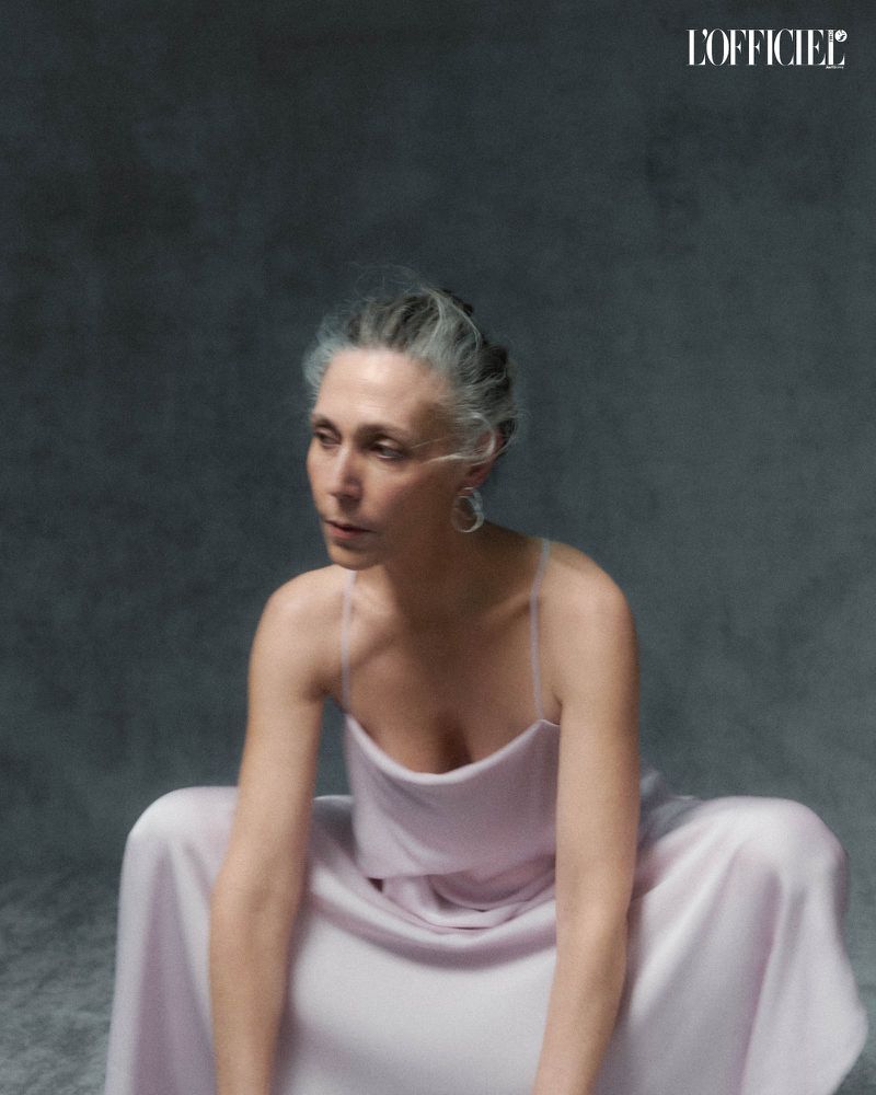 Elegant woman with silver hair poses in a light purple dress against a grey background, captured in a portrait fashion shot.