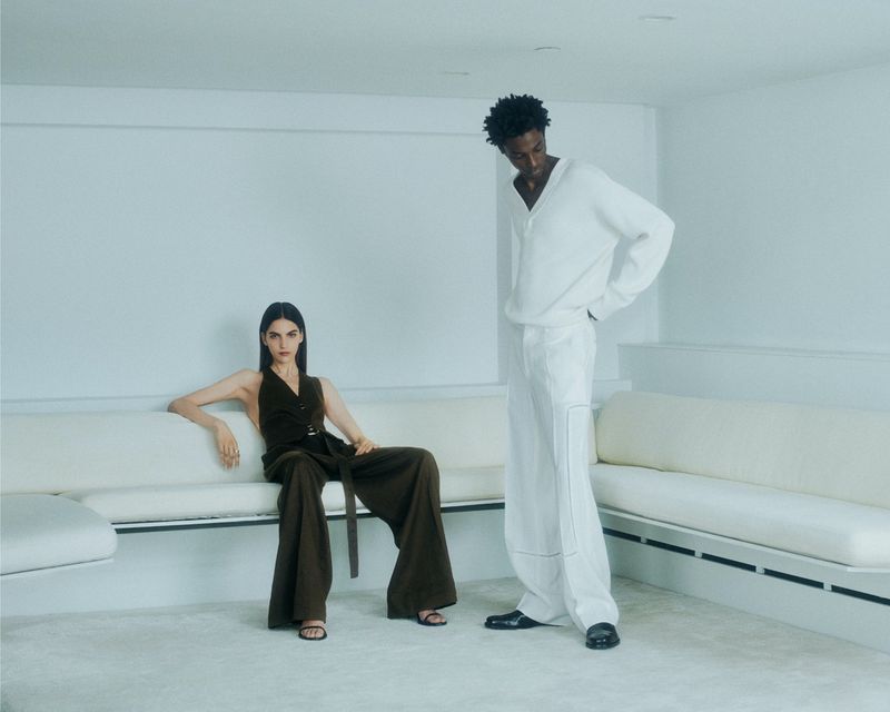Two fashion models pose indoors on modern couches against a white wall, one in an olive halter top with wide-leg pants, the other in white separates.