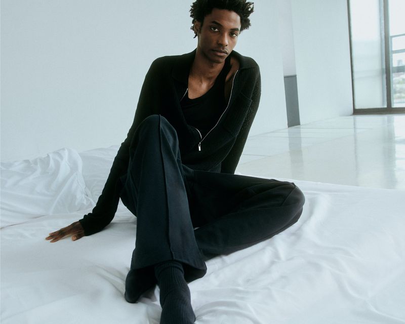 A Black male model poses in a relaxed seated position on a white bed with a window and wall background, wearing a dark zip-up jacket and pants.