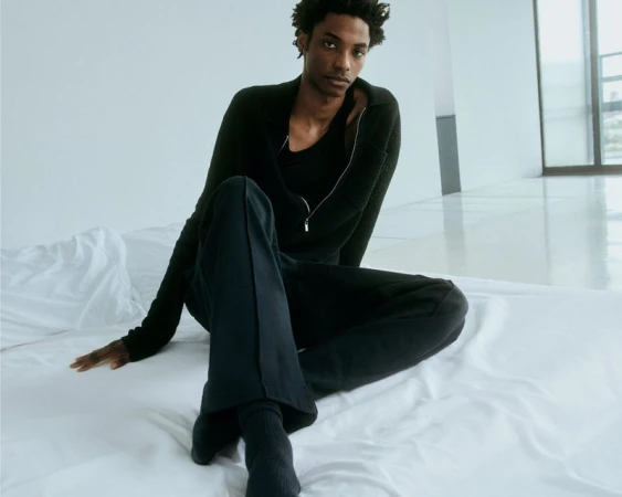 A Black male model poses in a relaxed seated position on a white bed with a window and wall background, wearing a dark zip-up jacket and pants.