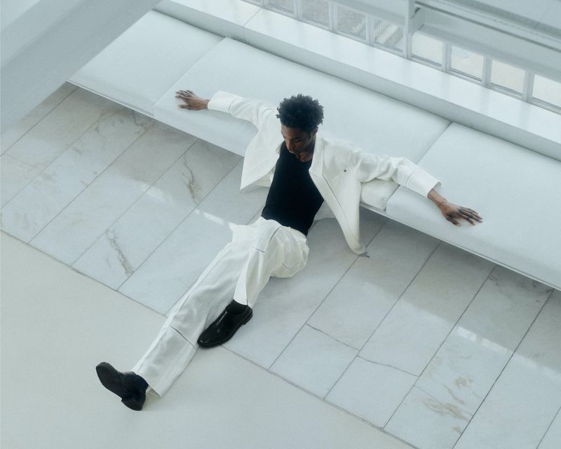 Overhead shot of a young Black male in white suit reclining on a white platform, wearing black top and shoes, in bright daylight.