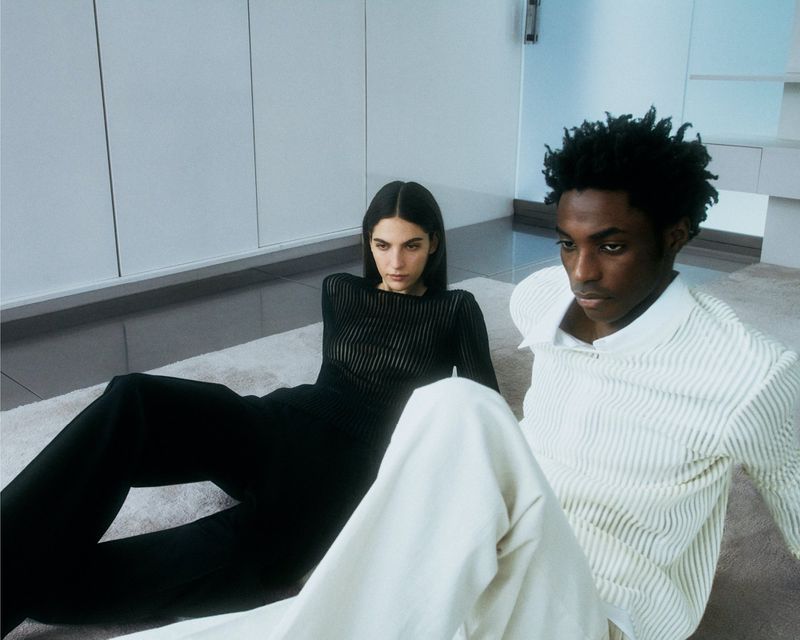 Two fashion models pose in an indoor setting on a cream carpet. A woman wears dark clothing; a man wears light-colored striped shirt and pants.