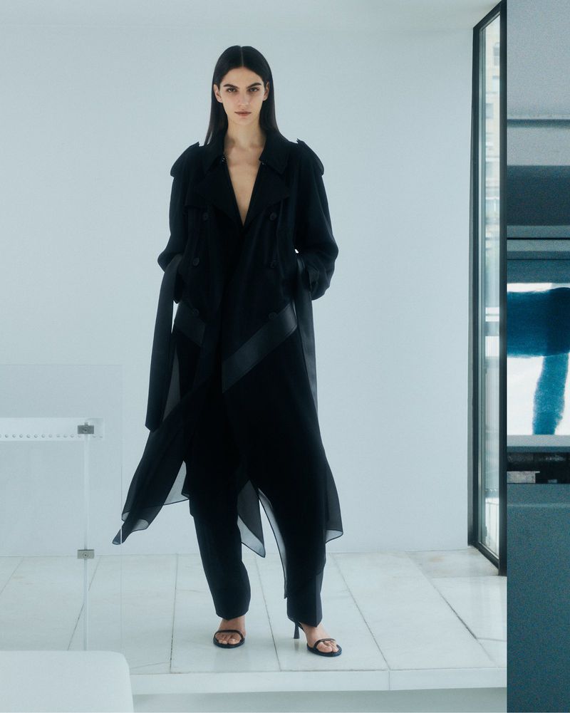 A stylish brunette woman in a long black trench coat and black heeled sandals poses in a minimalist white studio space.