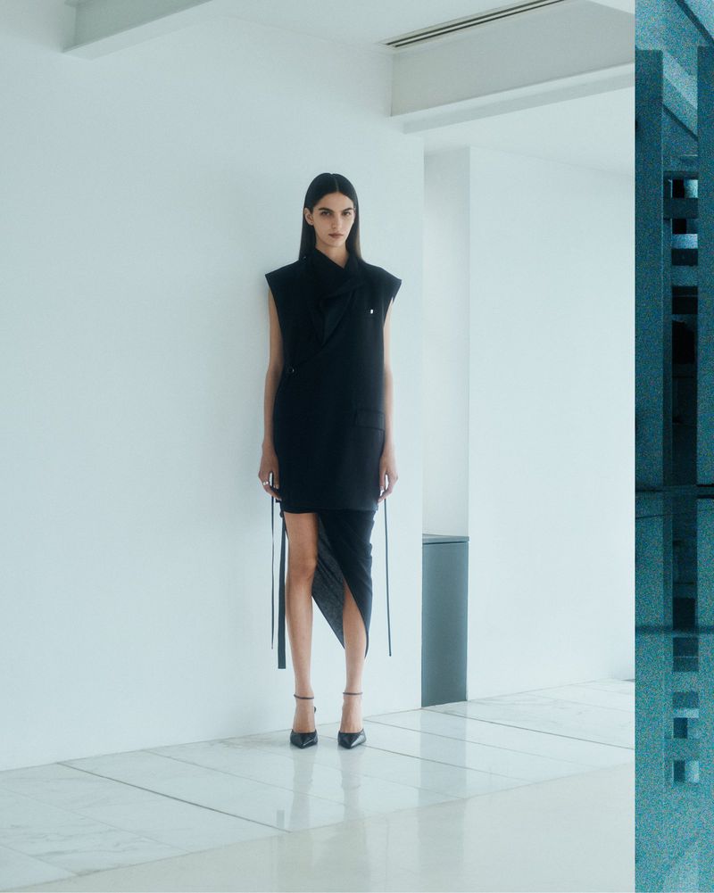 Fashion model in a sleeveless black dress and high heels poses indoors against a stark white wall and tiled floor with neutral light.