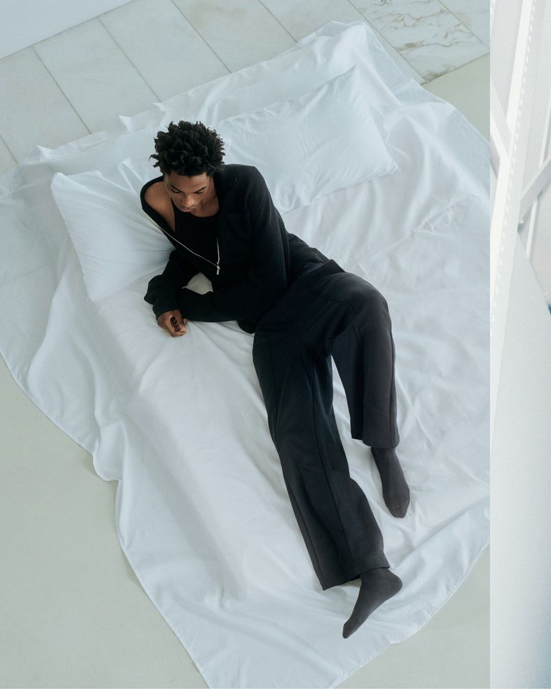 Overhead shot of a young adult man with dark skin and curly hair, lying on a white bed in black loungewear, in a brightly lit interior.