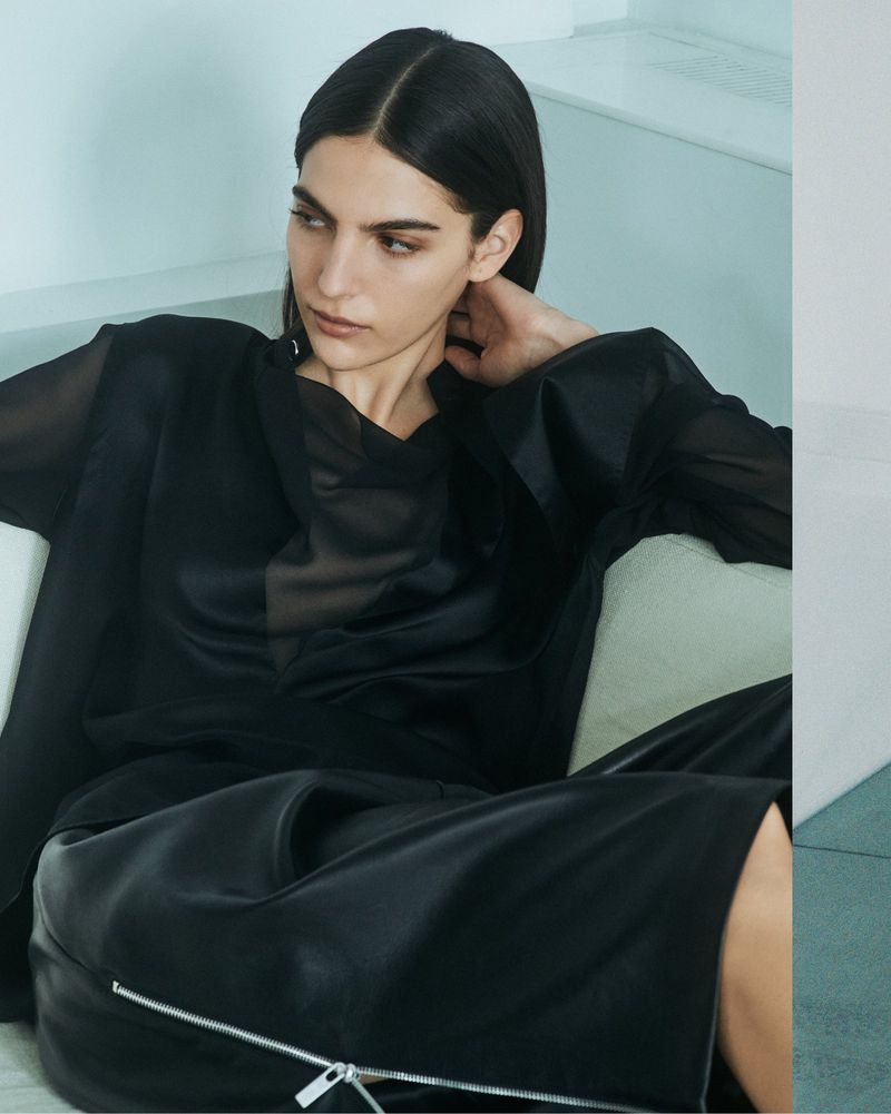 A woman in a sheer black top and leather pants reclines against a light background, looking off to the side in a fashion-forward pose.