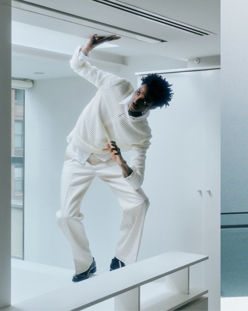 An African American male in a white textured sweater and pants strikes a pose on a white platform in a minimalist white room with large windows.