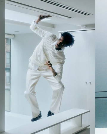 An African American male in a white textured sweater and pants strikes a pose on a white platform in a minimalist white room with large windows.