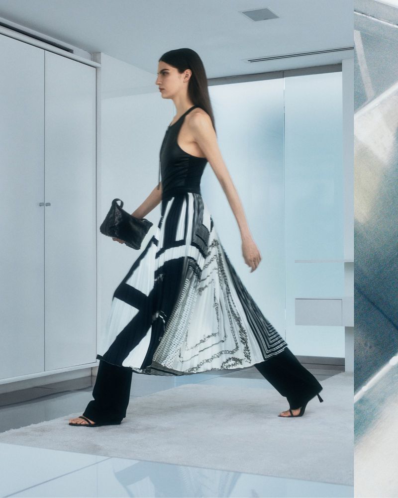 A woman walks in a minimalist indoor setting, wearing a black top, patterned white and black skirt, black pants, and carrying a handbag.