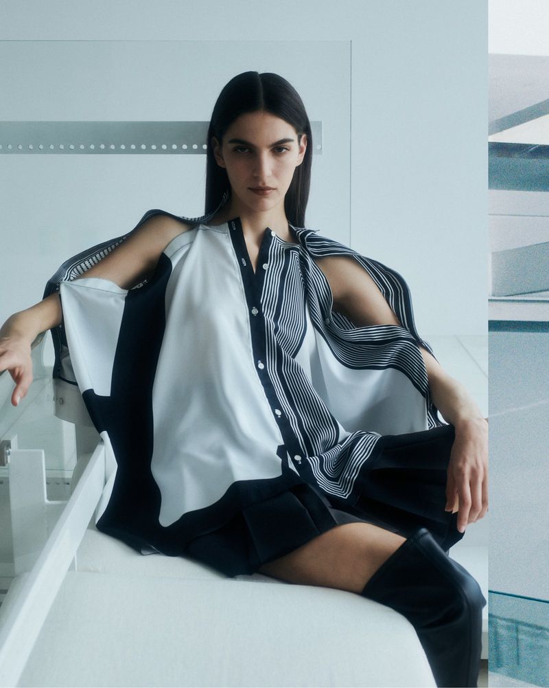 A brunette woman poses in a contemporary fashion shot. She wears a unique black and white top and black shorts, seated indoors.