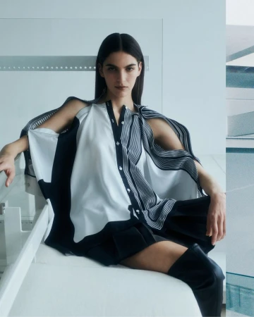 A brunette woman poses in a contemporary fashion shot. She wears a unique black and white top and black shorts, seated indoors.