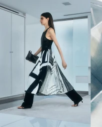 Model walking in a black top and geometric patterned skirt holding a black handbag in a modern interior