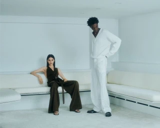 A woman in a dark brown jumpsuit seated beside a standing man dressed in an all-white ensemble in a minimalist room.