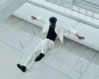 Model seated on a white tiled floor wearing a white suit jacket and trousers over a black tank top, photographed from an overhead angle.
