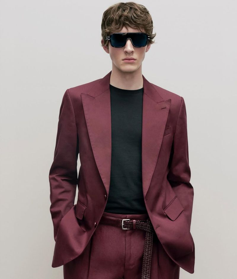 A stylish adult male poses indoors wearing sunglasses, a black t-shirt, and a burgundy suit with a braided belt, presenting a fashion-forward look.