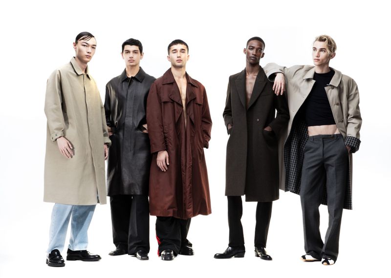 A group of five young adult male models wearing various fashionable overcoats in front of a bright white backdrop in a studio setting.
