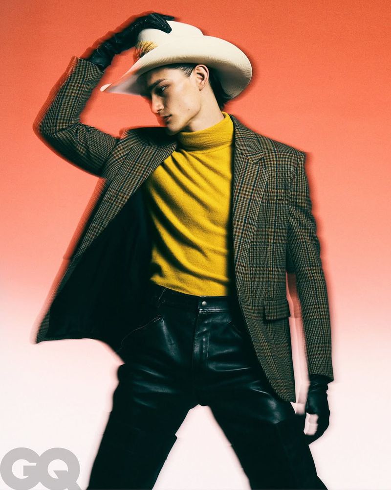 Fashion portrait of a young male adult in a yellow turtleneck, black leather pants, and a plaid blazer, topped with a white cowboy hat.