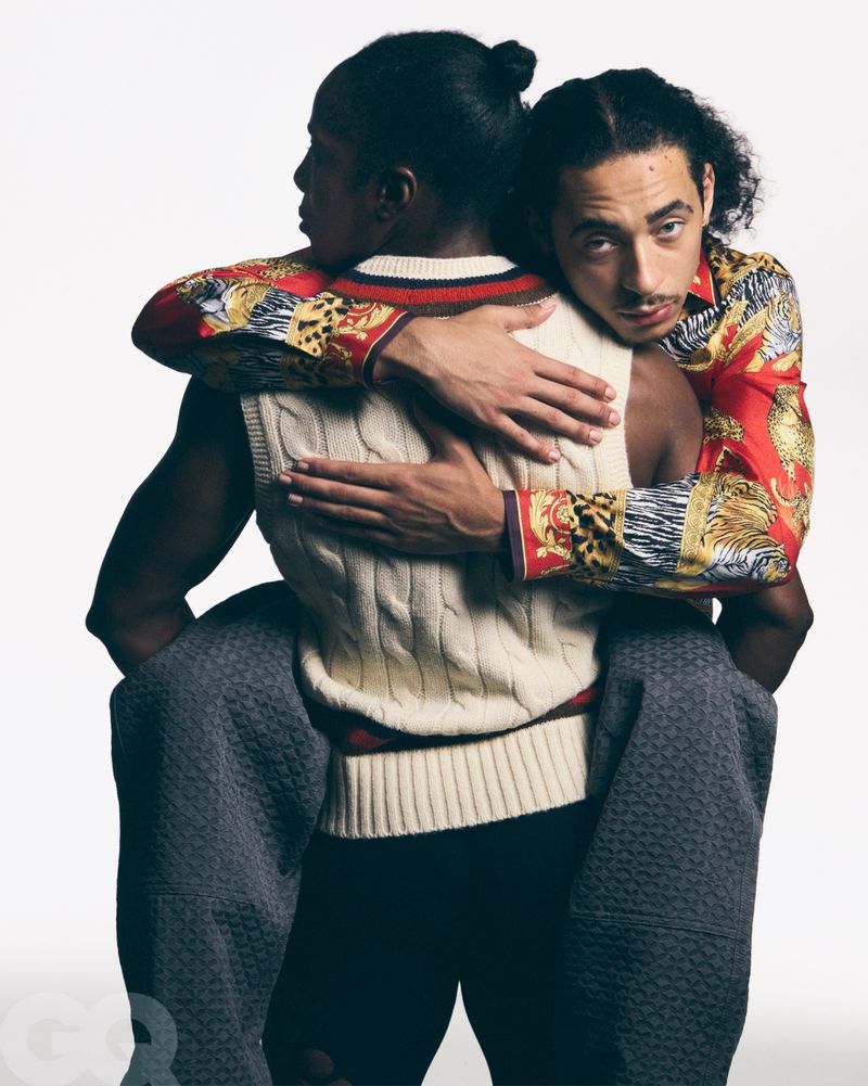 Two men in patterned shirts and a sweater vest embrace in a studio setting with a white background and soft lighting.