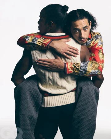 Two men in patterned shirts and a sweater vest embrace in a studio setting with a white background and soft lighting.