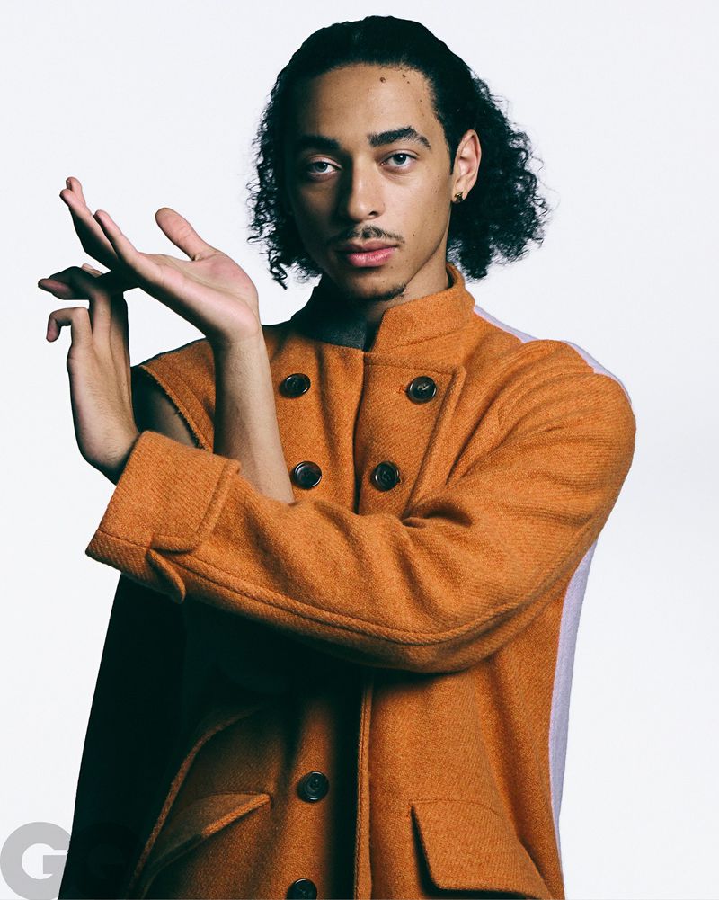 Fashion portrait of a young adult male with curly hair, wearing an orange button-up jacket, against a white background.