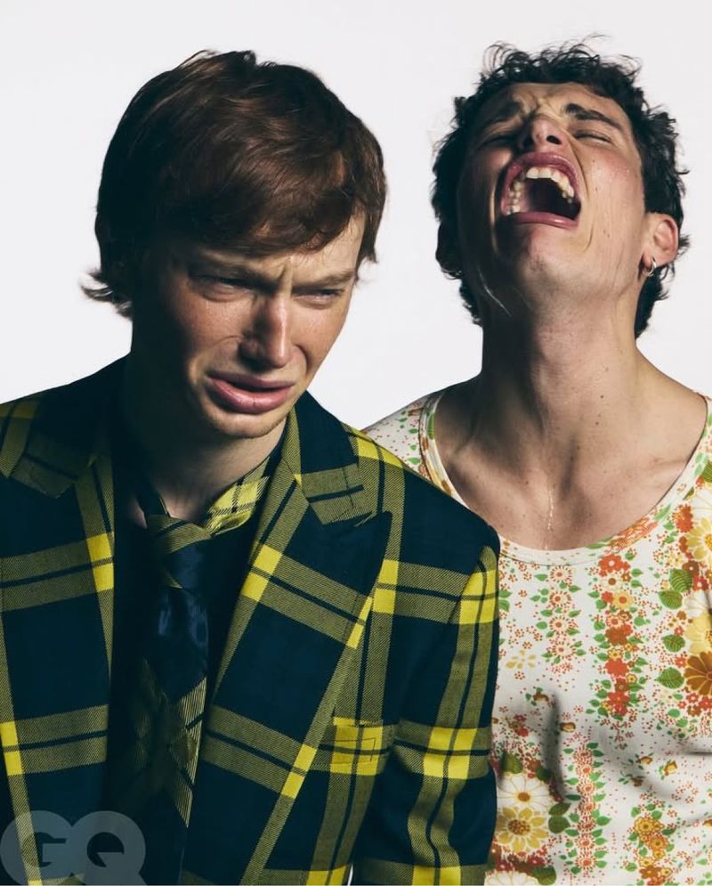 Two young adult men, one in plaid jacket looking upset, the other with mouth open and eyes closed, posing in front of a white background.