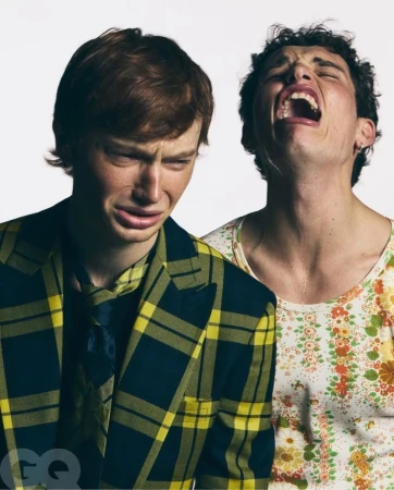 Two young adult men, one in plaid jacket looking upset, the other with mouth open and eyes closed, posing in front of a white background.