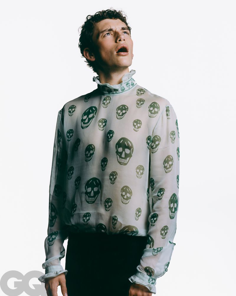 Fashionable adult male with open mouth and curly hair poses indoors wearing sheer skull-print top in a studio setting with bright lighting.