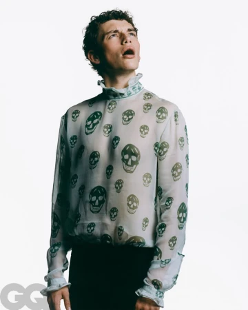 Fashionable adult male with open mouth and curly hair poses indoors wearing sheer skull-print top in a studio setting with bright lighting.