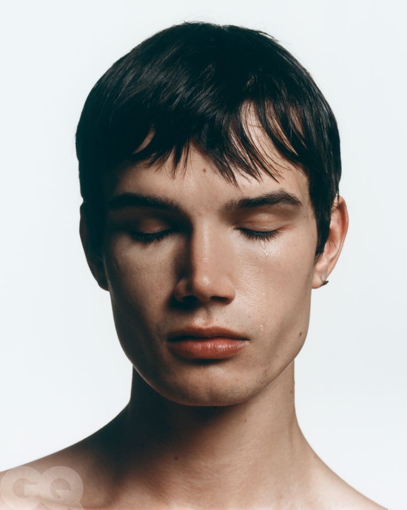 Close-up studio portrait of a young adult male with dark hair, eyes closed, and a visible tear. Against a clean white backdrop.