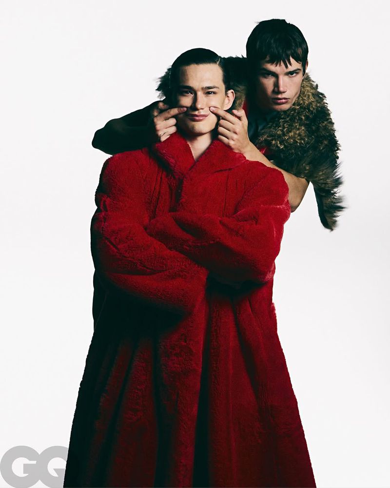 Two male models pose in a photo studio: one in a red faux fur coat, the other in a fur shawl, fingers at smile mimicking a happy face.