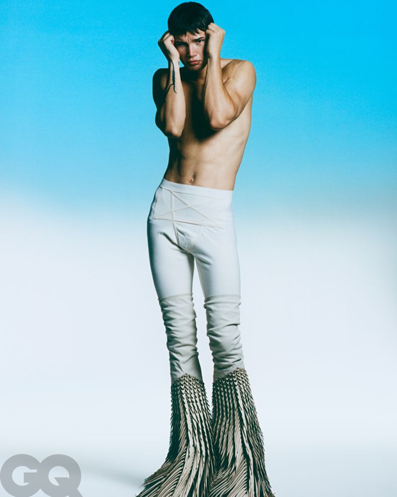 Shirtless male model in white fringed pants strikes a pose against a vibrant blue gradient backdrop in a fashion editorial shot.