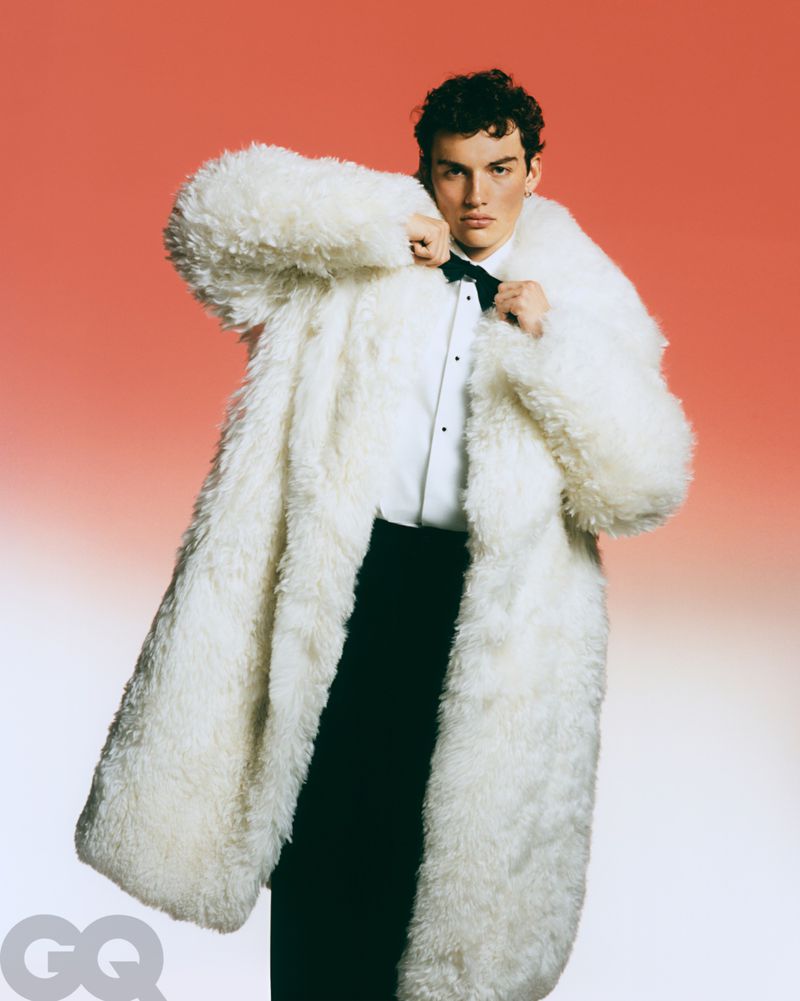Young man in a white, oversized faux fur coat, white collared shirt, and black bow tie and pants, posing against a gradient red background.