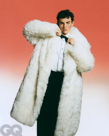 Young man in a white, oversized faux fur coat, white collared shirt, and black bow tie and pants, posing against a gradient red background.