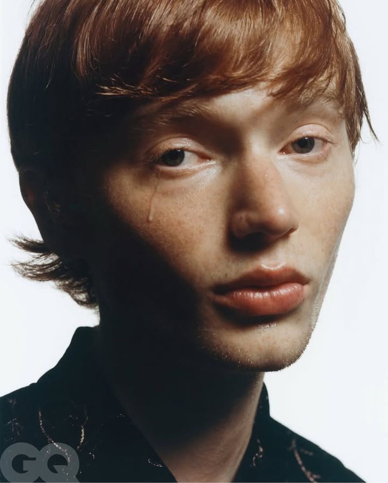 Close-up portrait of a young adult male with red hair and tear on cheek, posing indoors against a white background, in a fashion shot.