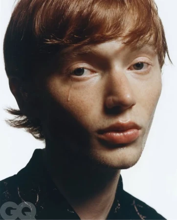 Close-up portrait of a young adult male with red hair and tear on cheek, posing indoors against a white background, in a fashion shot.