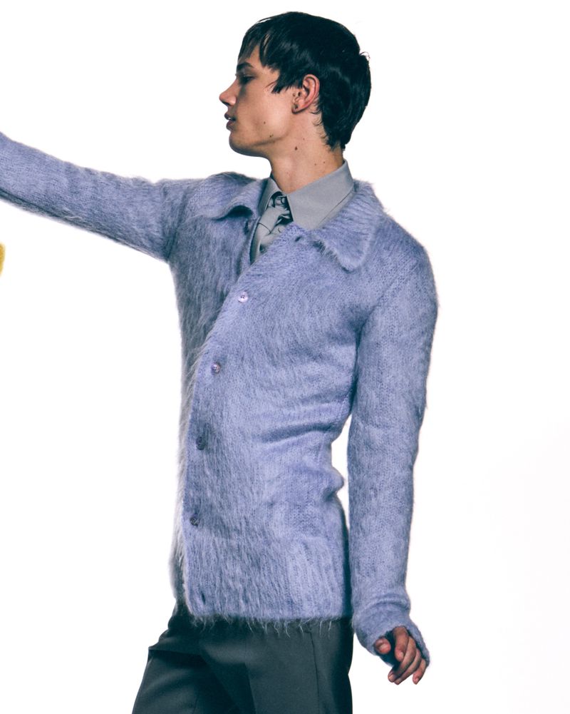 A light-skinned male model wearing a blue cardigan, grey shirt and tie, and grey pants, posing in a bright indoor photo studio.
